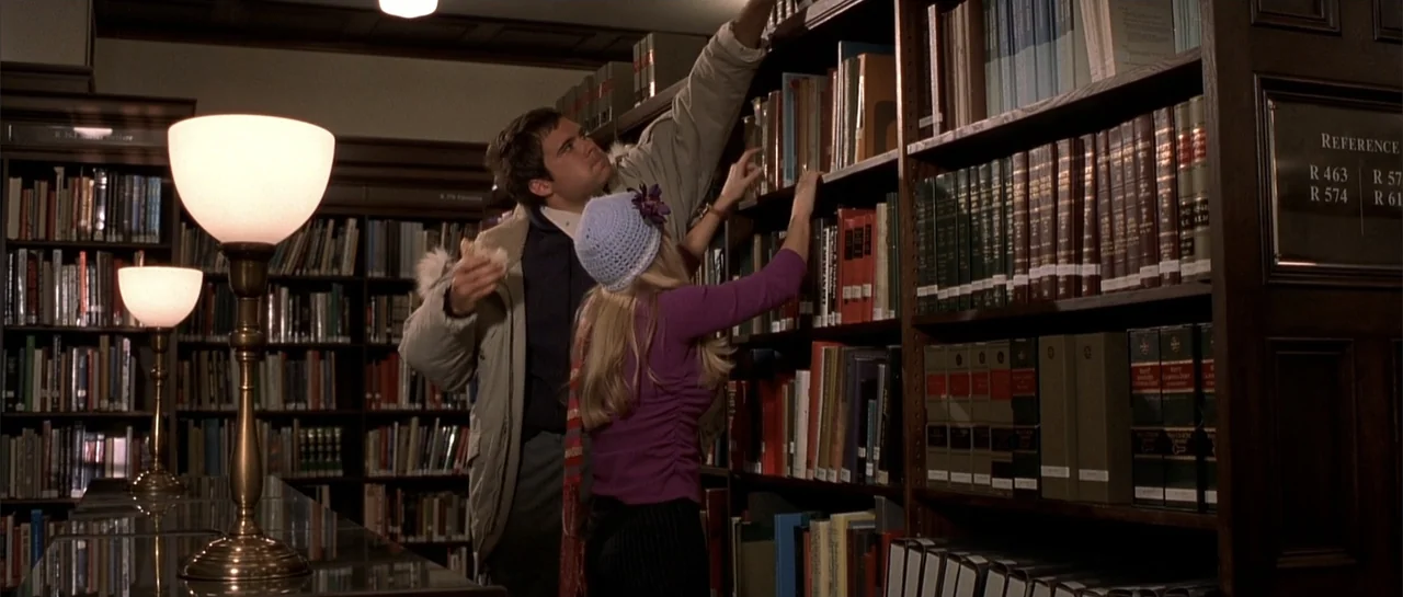 Legally Blonde (2001) movie still: medium — A quiet library interior features dark wood shelves filled with numerous reference books. A man and a…