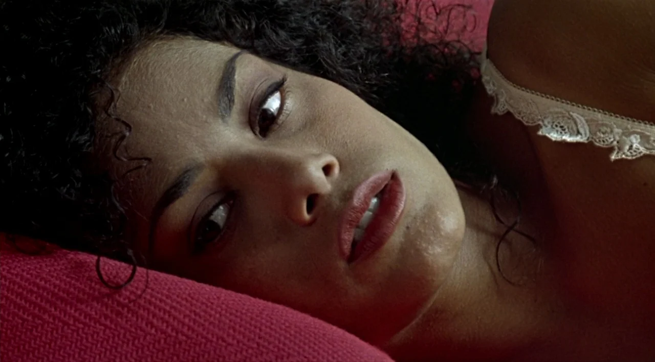 Kika (1993) movie still: closeup — A person lies on a soft surface with bright red fabric and white lace. The subject gazes upward with eyes focused…