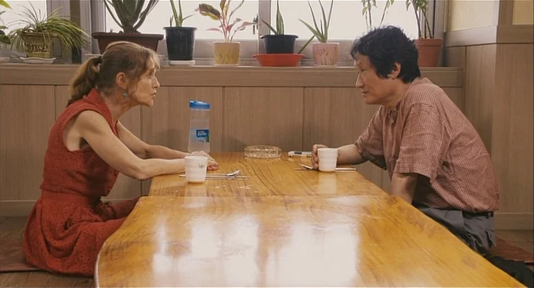 In Another Country (2012) movie still: medium — A wooden table in a brightly lit room with a row of potted plants. Two people are sitting opposite…