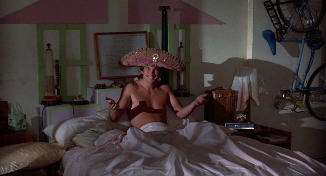 Breathless (1983) movie still: medium — A bedroom contains white bed sheets, a wall-mounted bicycle, and pink-green wall paneling. A man sits…