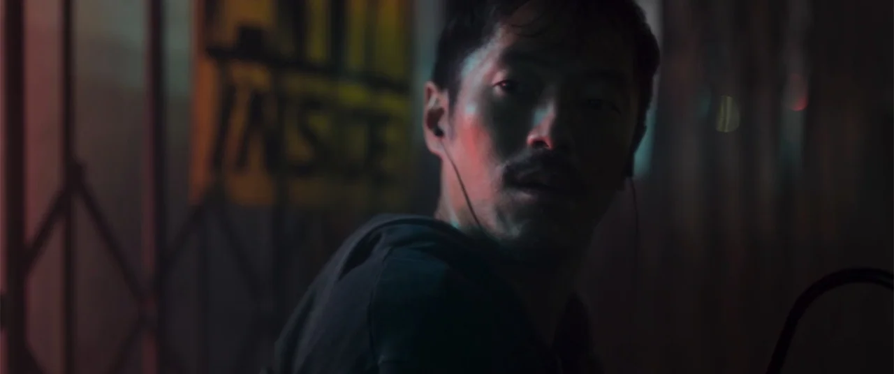 Phobias (2021) movie still: closeup — A man stands outdoors at night near a dark metal gate. He looks over his shoulder while wearing dark clothing…