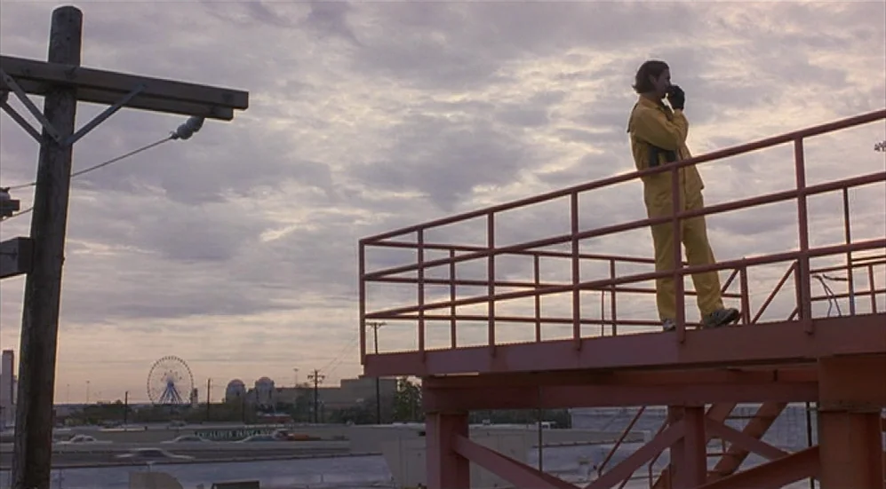 Bottle Rocket (1996) movie still: wide — An industrial metal scaffolding platform overlooks a distant city skyline with a ferris wheel. A man in a…