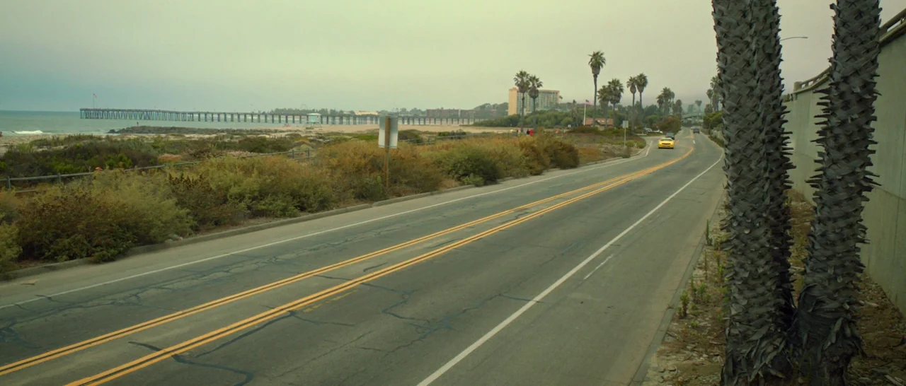 God Bless America (2011) movie still: wide — An asphalt coastal road runs parallel to a sand beach and wooden pier. A yellow car travels on the road…
