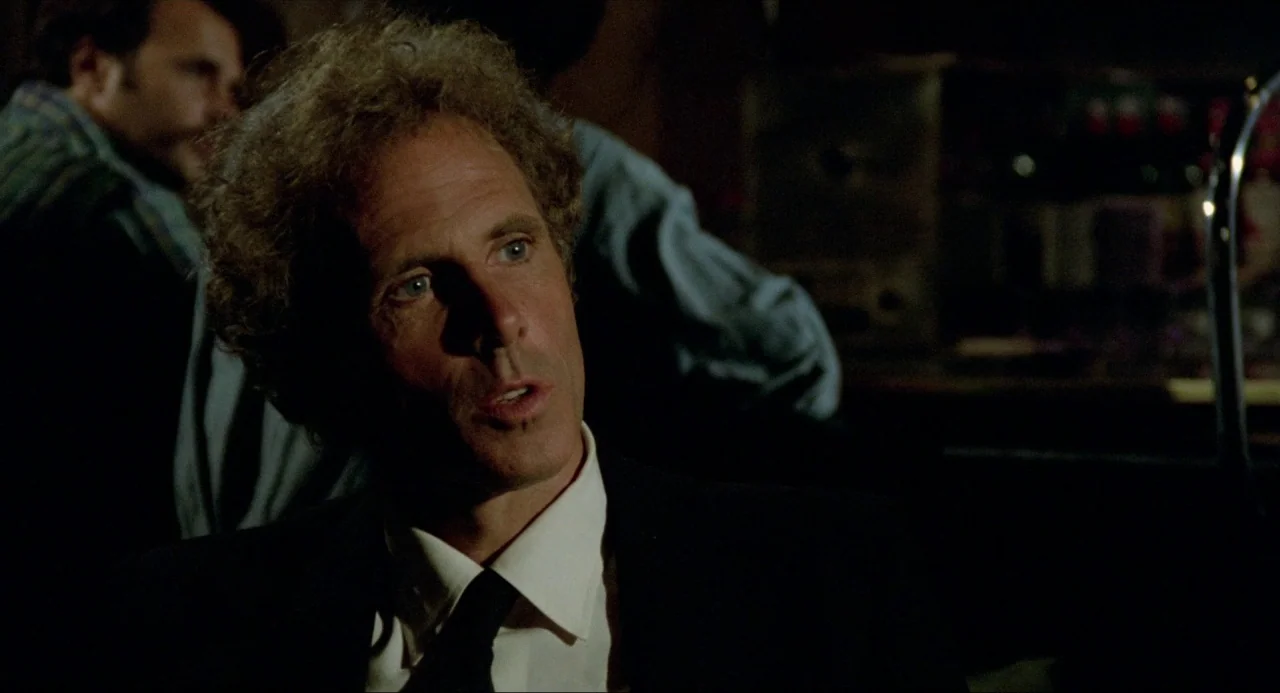The Driver (1978) movie still: closeup — A dimly lit bar setting with blurred figures in the background of the frame. A man in a suit jacket turns…
