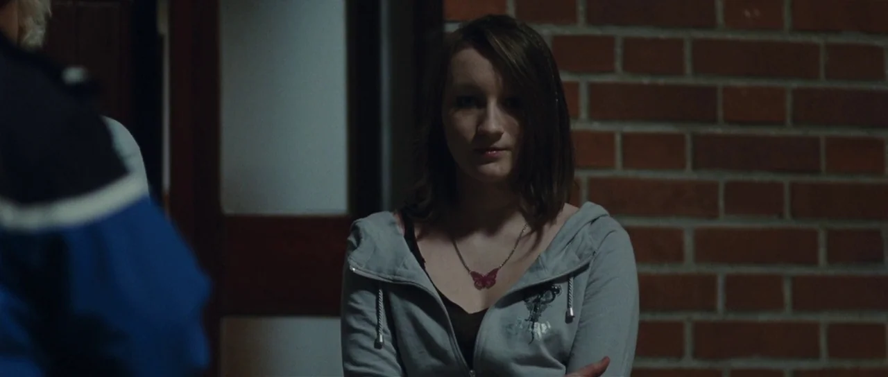 Our Day Will Come (2010) movie still: medium — A dark outdoor space beside a red brick wall at night. A young woman in a grey hoodie stands facing…
