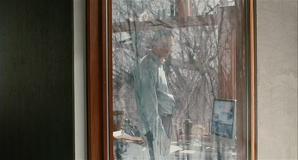 Chloe (2009) movie still: medium — A man stands inside a room viewed through a large windowpane. Outside, bare trees are visible while the man looks…
