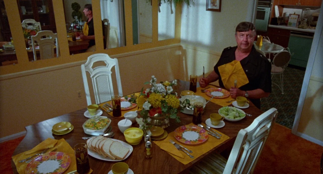 Polyester (1981) movie still: medium — A dining room decorated with yellow walls and floral patterned plates on the table. An older man wearing a…