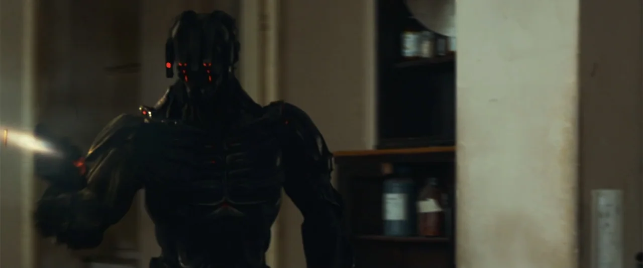 The Condemned 2 (2015) movie still: medium — A dimly lit indoor space with blurred storage units in the background. A dark, armored robotic entity…