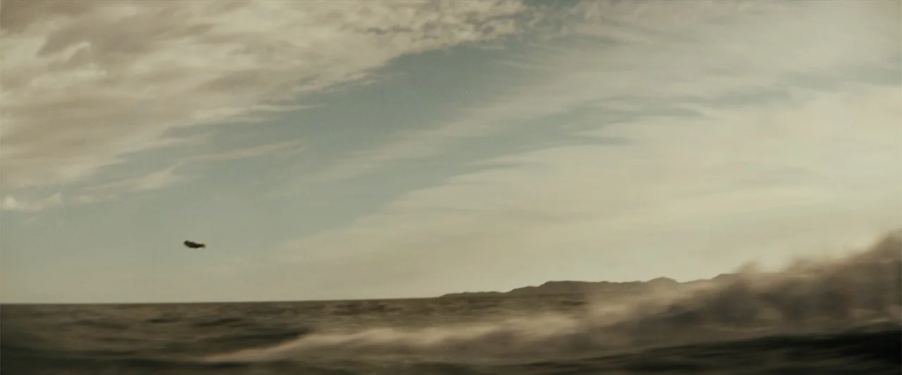 Black Adam (2022) movie still: establishing — An open expanse of ocean with a distant coastline visible beneath a cloudy sky. A small object moves…
