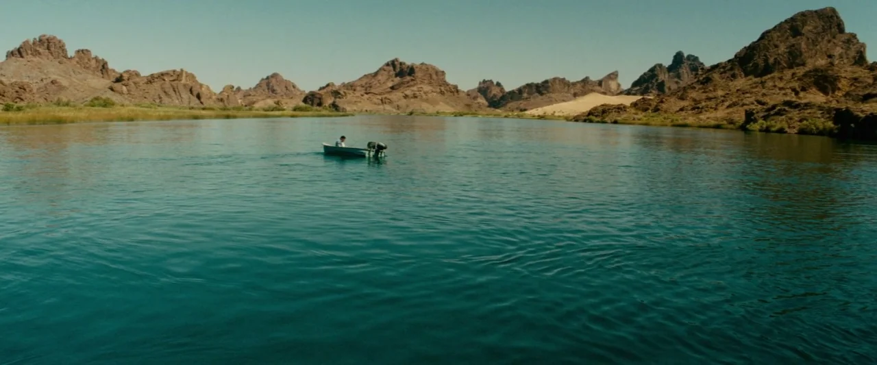 Piranha 3D (2010) movie still: wide — A wide, calm river flows between dry, rugged desert mountains under a clear sky. A lone person operates a…