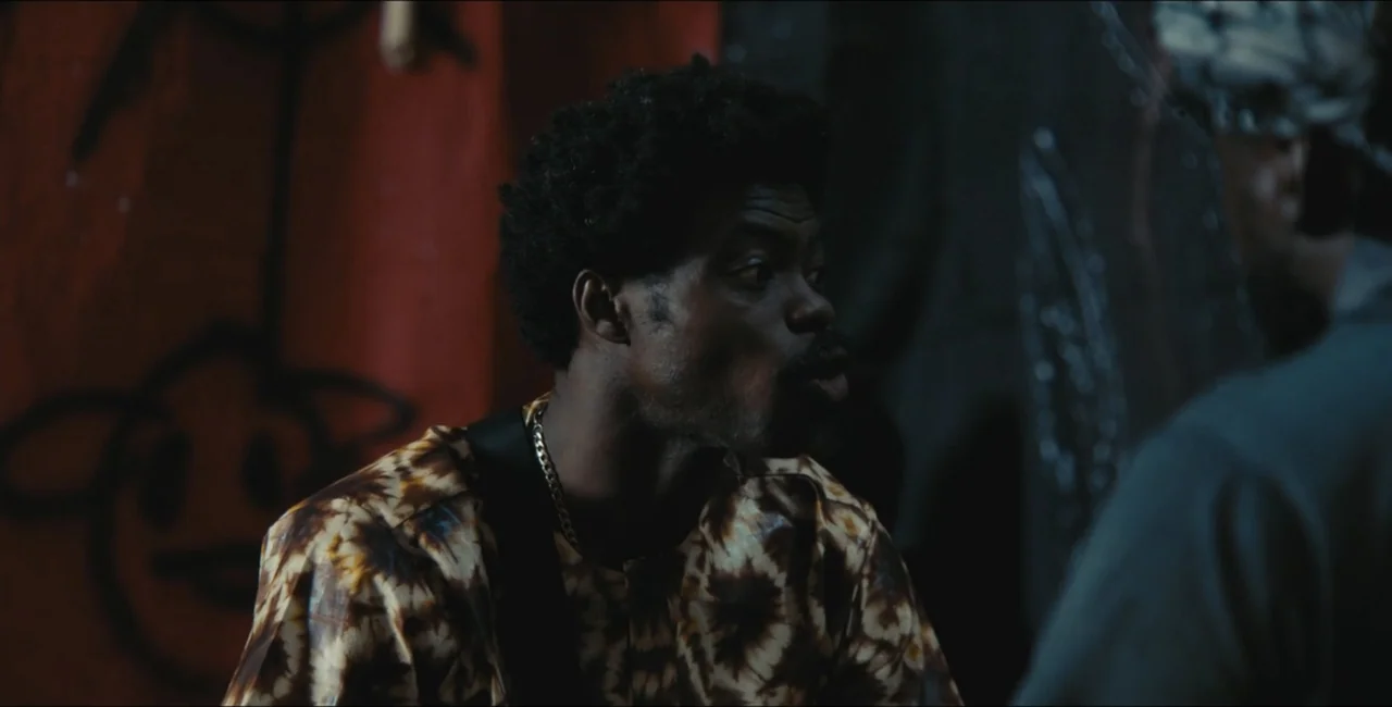 Project Assault (2025) movie still: medium — A dimly lit room with dark walls features graffiti art in the background. A man with an afro and…