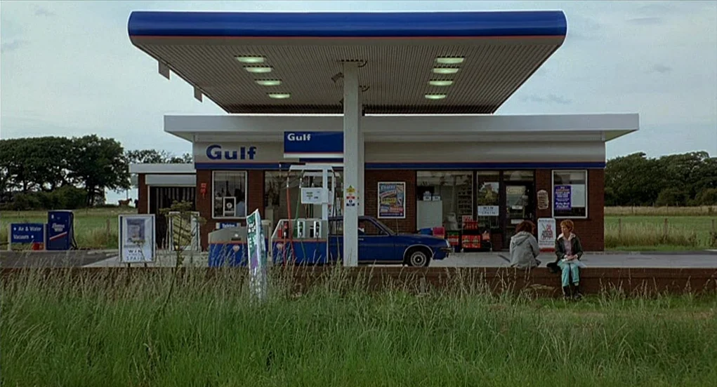 Butterfly Kiss (1995) movie still: wide — A rural Gulf gas station stands under an overcast sky surrounded by green fields. Two women sit on the…