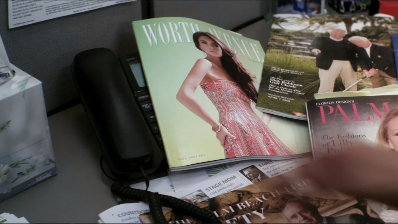 Parker (2013) movie still: closeup — A cluttered office desk surface features multiple glossy magazines and a black landline phone. A hand reaches…