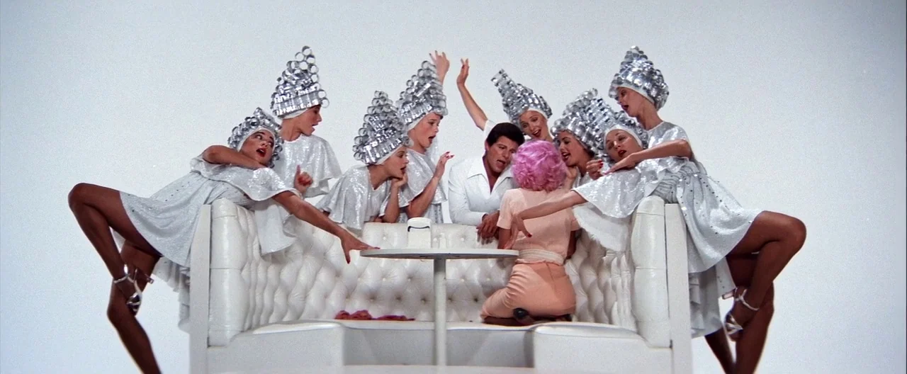 Grease (1978) movie still: wide — A white diner booth sits isolated against a pure white background. Several women wearing silver hair rollers and…