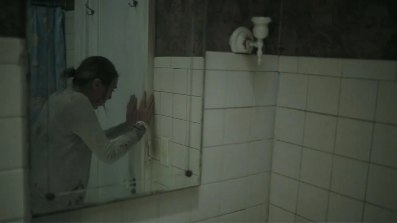 Silent House (2011) movie still: medium — A tiled bathroom with patterned wallpaper and a mirror mounted on the wall. A young woman with disheveled…