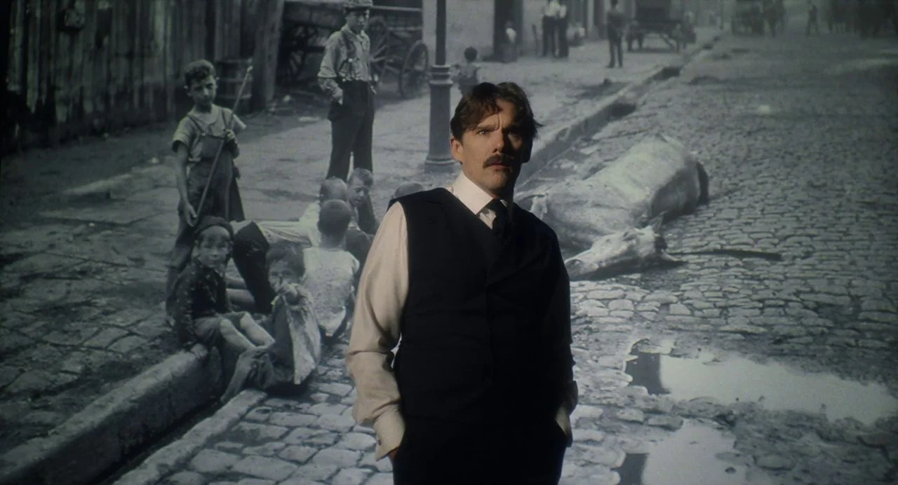 Tesla (2020) movie still: medium — A man stands in front of a large monochrome projection of a cobblestone street. The backdrop shows historical…