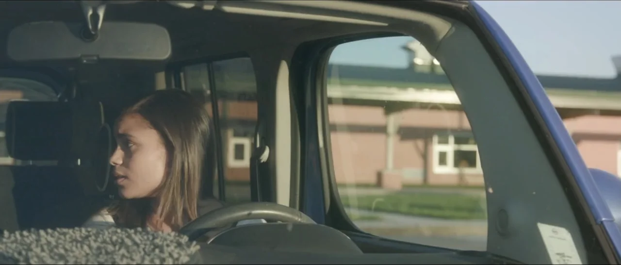 If You're Gone (2019) movie still: medium — A teenager sits in the driver's seat of a car during the day. She looks out the side window at a blurry…