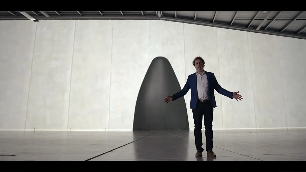 Wild Wild Space (2024) movie still: wide — A man stands in an expansive white industrial hangar with a large arched opening. He stands in the center…