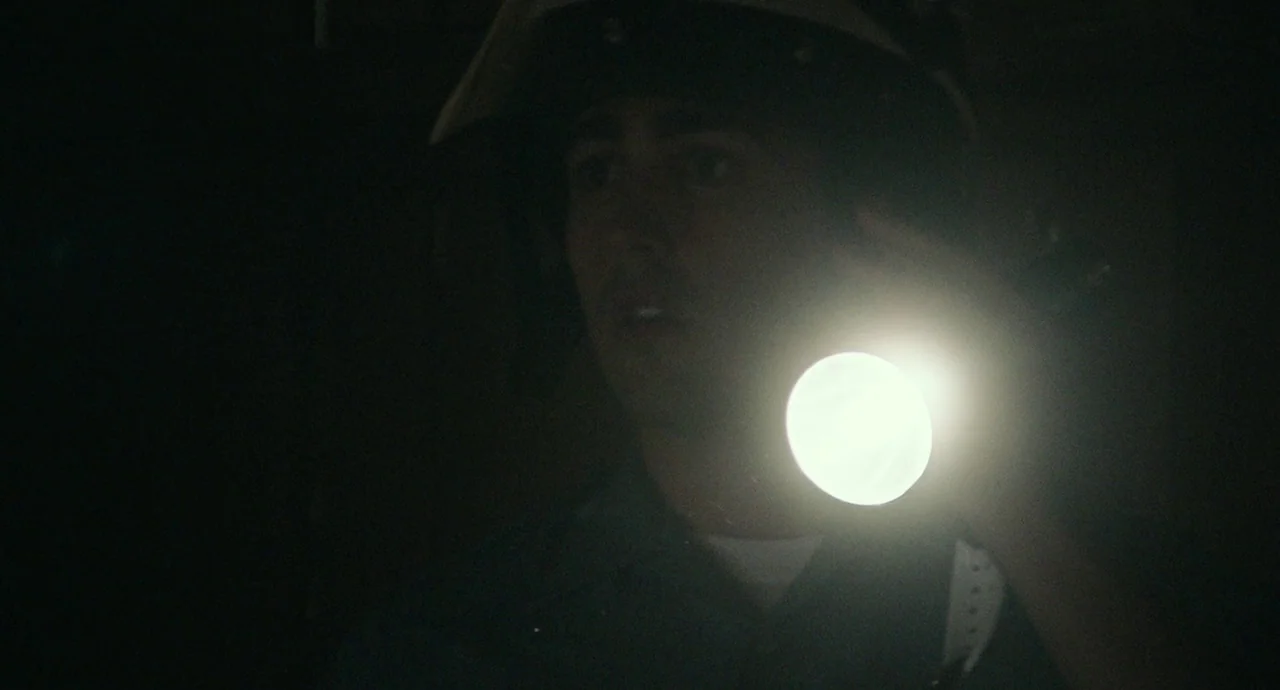 Detroit (2017) movie still: closeup — A dark, obscured space is illuminated by a single handheld light source. A young man wearing a helmet holds a…