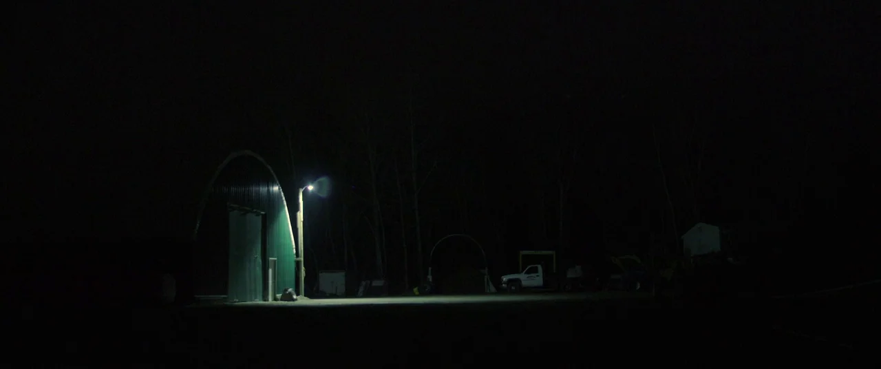 The Void (2016) movie still: wide — A nighttime industrial area features a corrugated metal structure illuminated by a single lamp. A pickup truck…