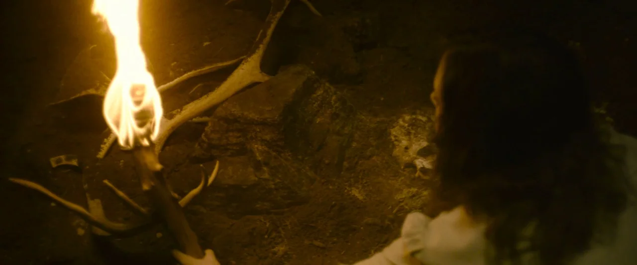 Wildling (2018) movie still: medium — A dark cave interior with scattered animal antlers resting on a dirt floor. A person holds a lit torch while…