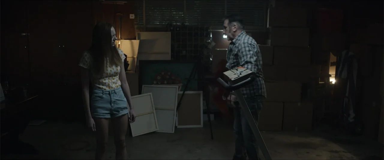 Captive (2020) movie still: medium — A cluttered storage room filled with cardboard boxes and canvas paintings in the dark. A man and a woman stand…