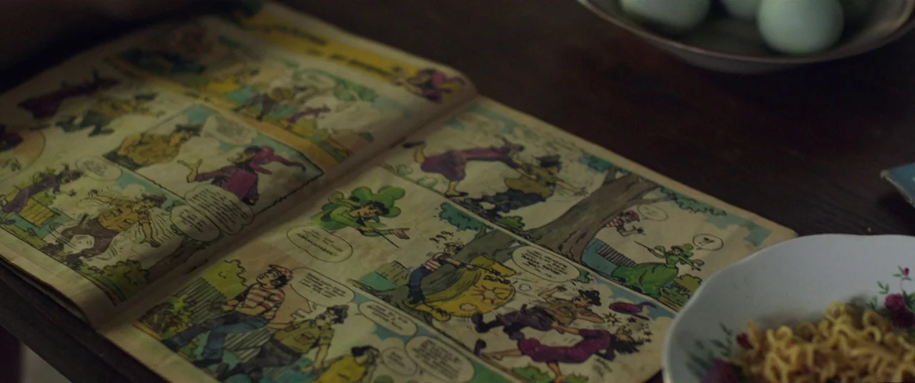 Satan’s Slaves (2017) movie still: closeup — A vintage comic book is spread open on a dark wood table. A bowl of noodles and a plate of eggs sit…