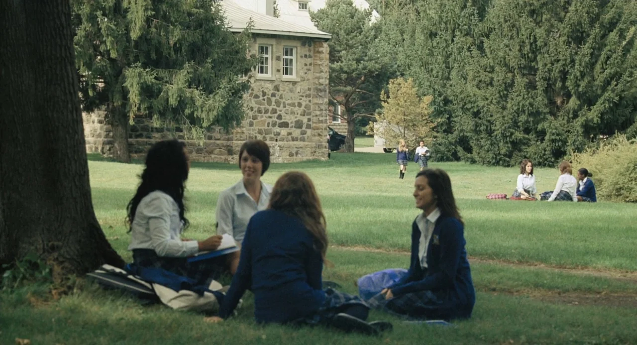 The Moth Diaries (2011) movie still: wide — A group of female students in school uniforms sit on the grass outside. Stone buildings and evergreen…