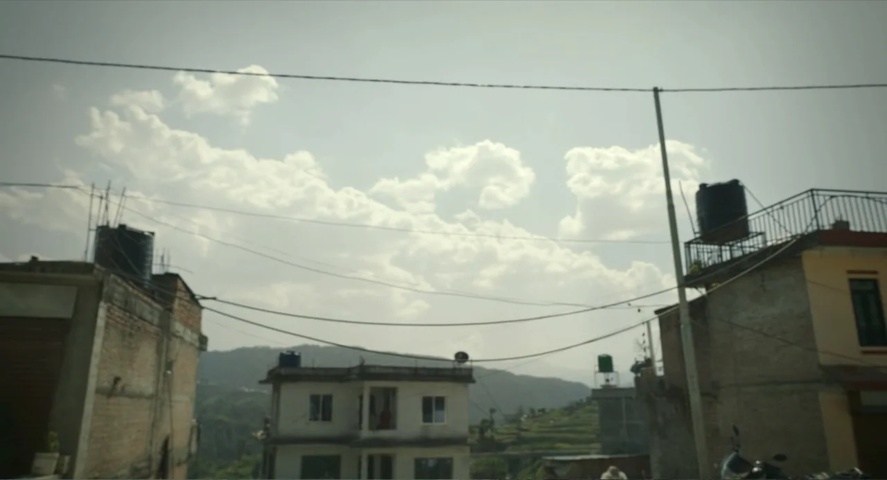 Ice Road: Vengeance (2025) movie still: establishing — An outdoor neighborhood scene shows residential buildings with water tanks under an overcast…
