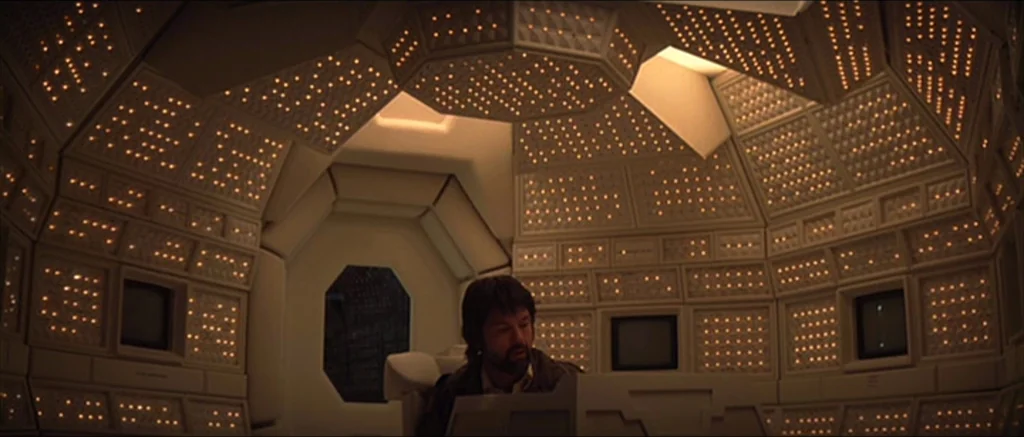 Alien (1979) movie still: medium — A futuristic compartment is covered in walls of repetitive grid-like backlit light panels. A man sits at a…