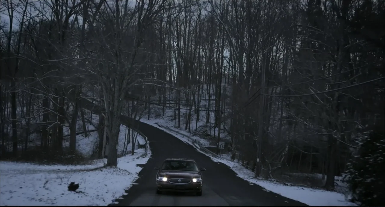 Freedom (2024) movie still: wide — A dark sedan travels down a winding, snow-covered road lined with leafless winter trees. The vehicle moves toward…