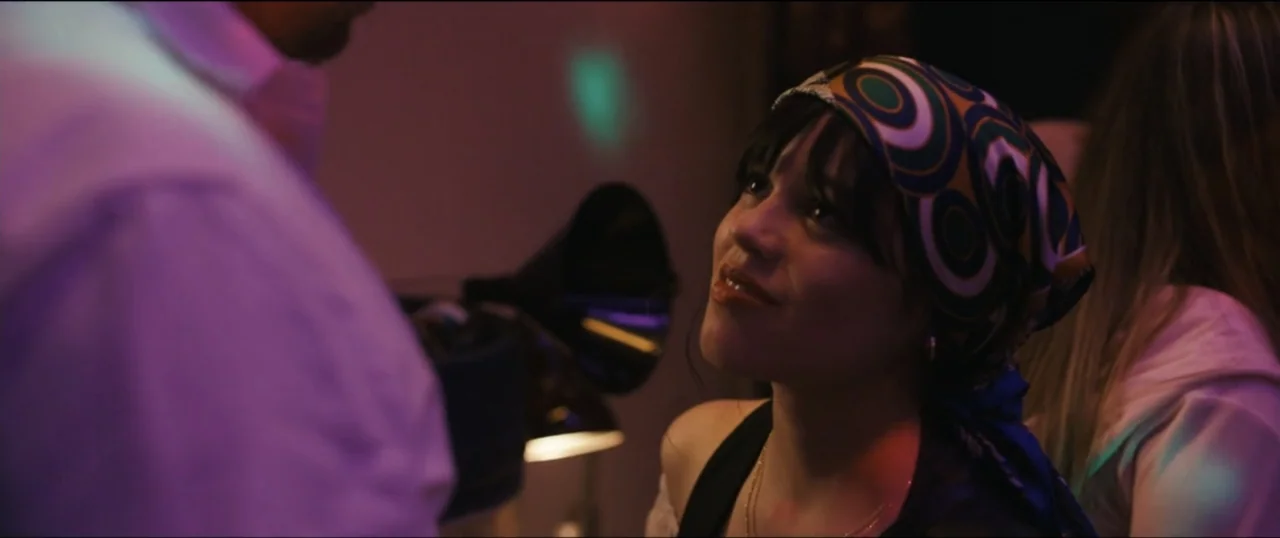 Scream VI (2023) movie still: medium — A dark indoor party setting with colorful ambient light reflected on dark surfaces. A woman wearing a…