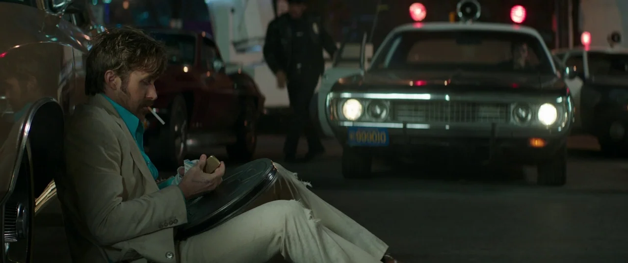 The Nice Guys (2016) movie still: medium — A dark, wet asphalt lot filled with vintage vehicles at night. A man in a light suit rests against a car…