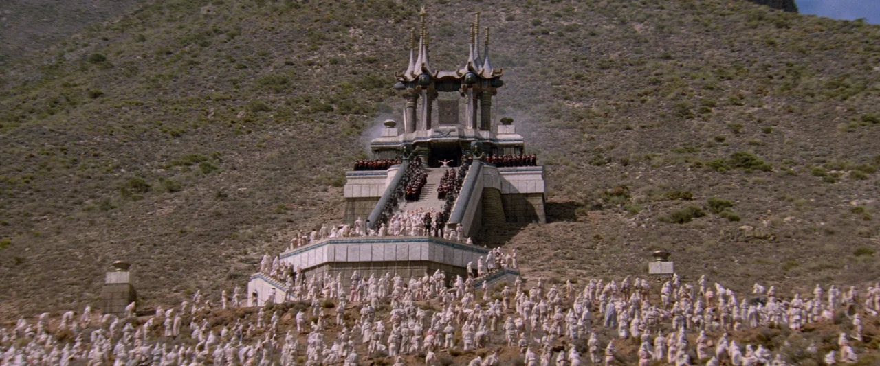 Conan The Barbarian (1982) movie still: establishing — A stone temple with tall spires stands on a rocky, scrub-covered mountain slope. A large…