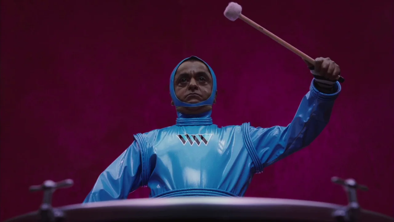 Charlie and The Chocolate Factory (2005) movie still: medium — A man in a bright blue costume stands before a large drum against magenta. He holds a…