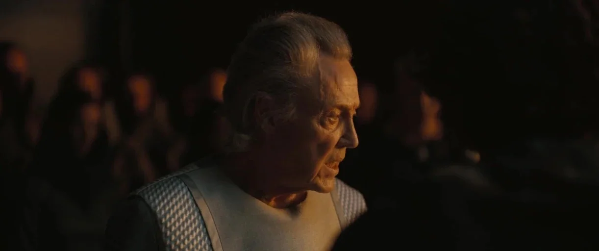 Dune: Part 2 (2024) movie still: closeup — A dark, dimly lit interior space with vague, out-of-focus figures in the background. An elderly man in a…