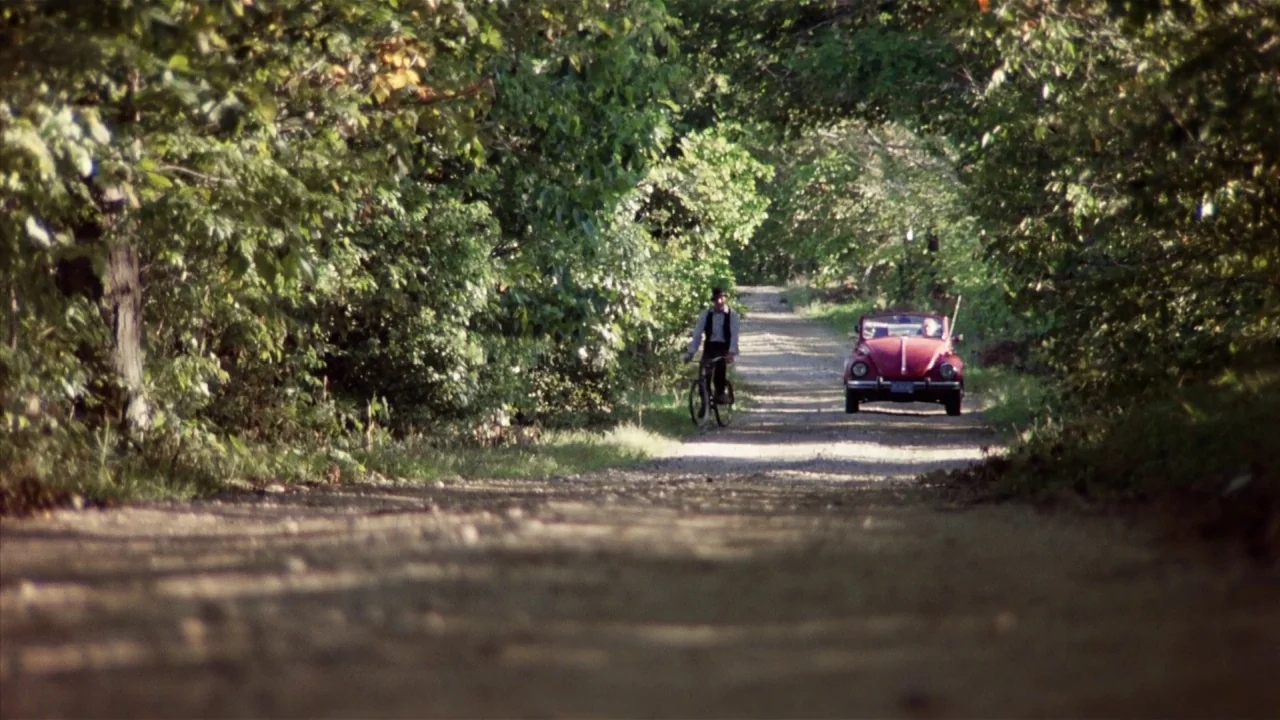 Friday the 13th Part 2 (1981) movie still: wide — A narrow dirt road is lined with dense deciduous trees forming a tunnel canopy. A man rides a…