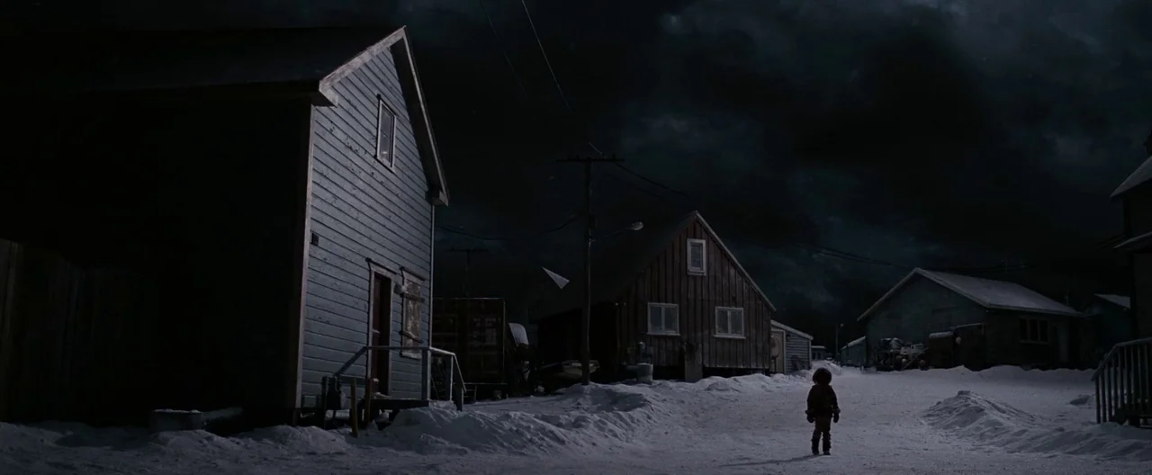 30 Days of Night (2007) movie still: wide — A dark, snow-covered village is surrounded by wooden buildings under a heavy storm sky. A small child…