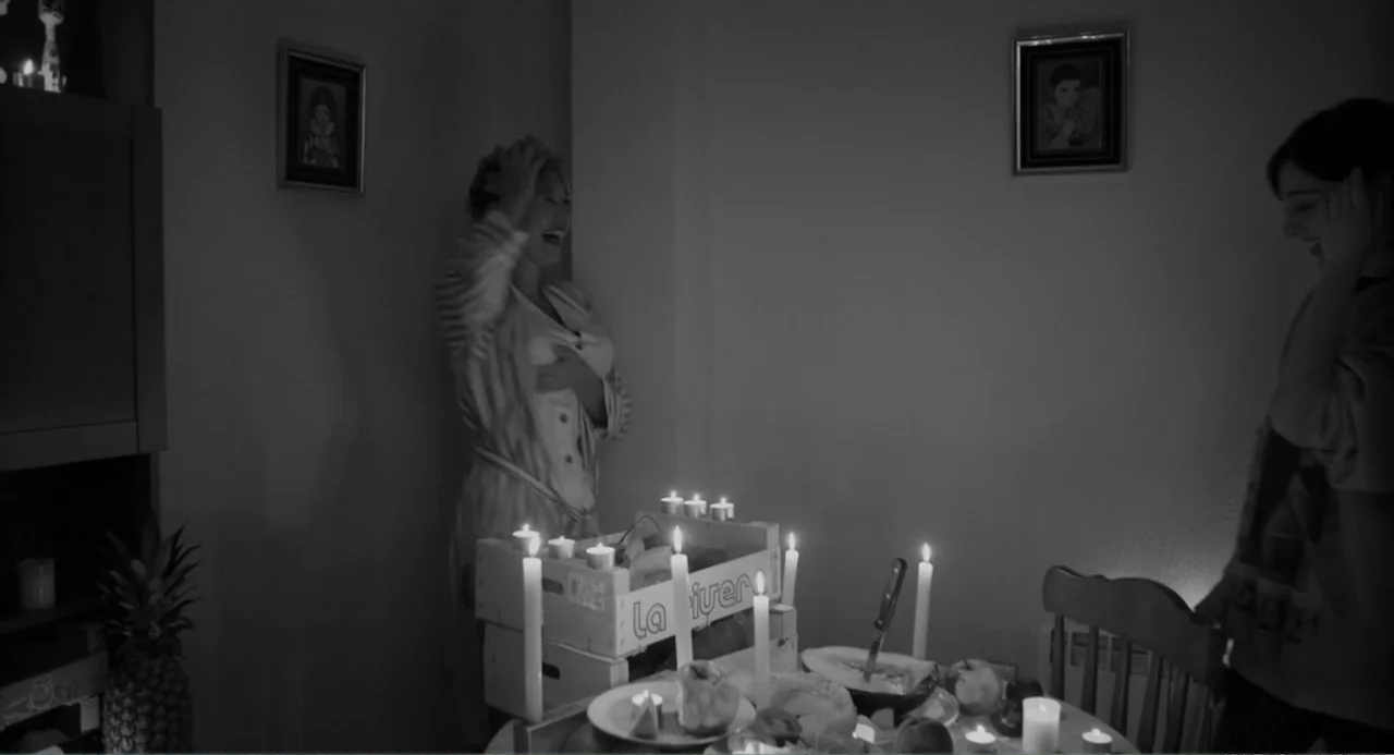 El Planeta (2021) movie still: medium — A dimly lit room features a table cluttered with candles and halved fruit. Two women stand on opposite sides…