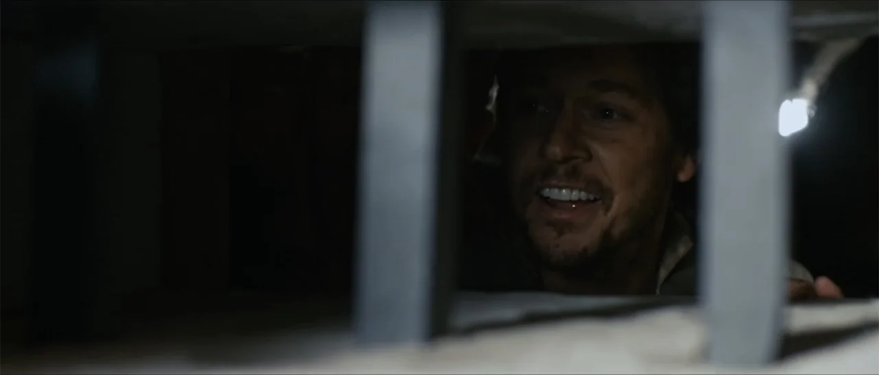 Out of Liberty (2019) movie still: closeup — A dark space defined by vertical metal bars blocking the view. A man stares through the gaps with wide…