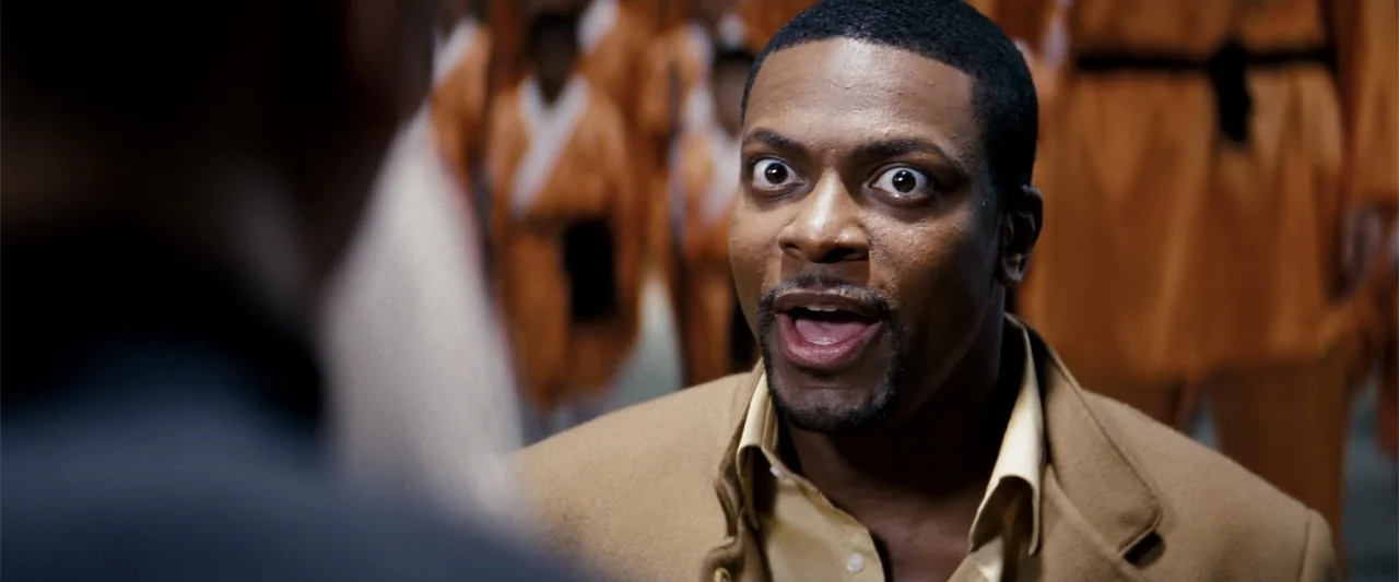 Rush Hour 3 (2007) movie still: closeup — An interior space features a foreground man in a tan jacket facing away from camera. Behind him, a group…