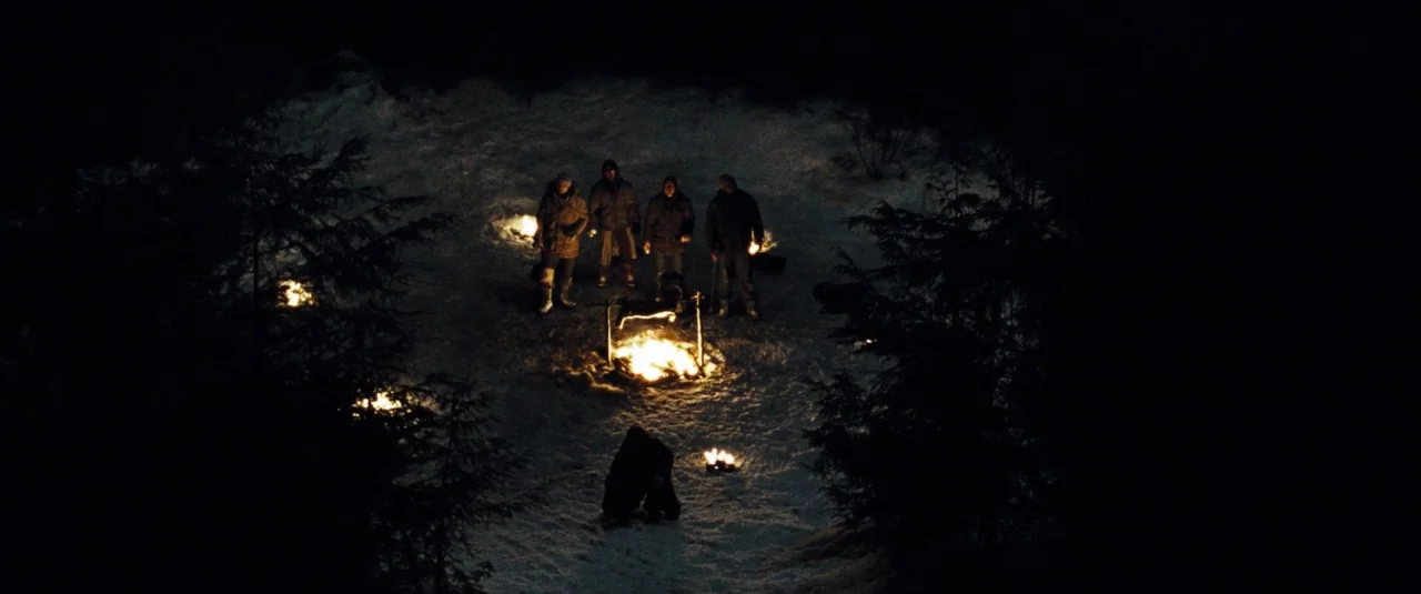 The Grey (2012) movie still: wide — A dark, snow-covered clearing surrounded by tall coniferous trees is illuminated by small fires. Five people…