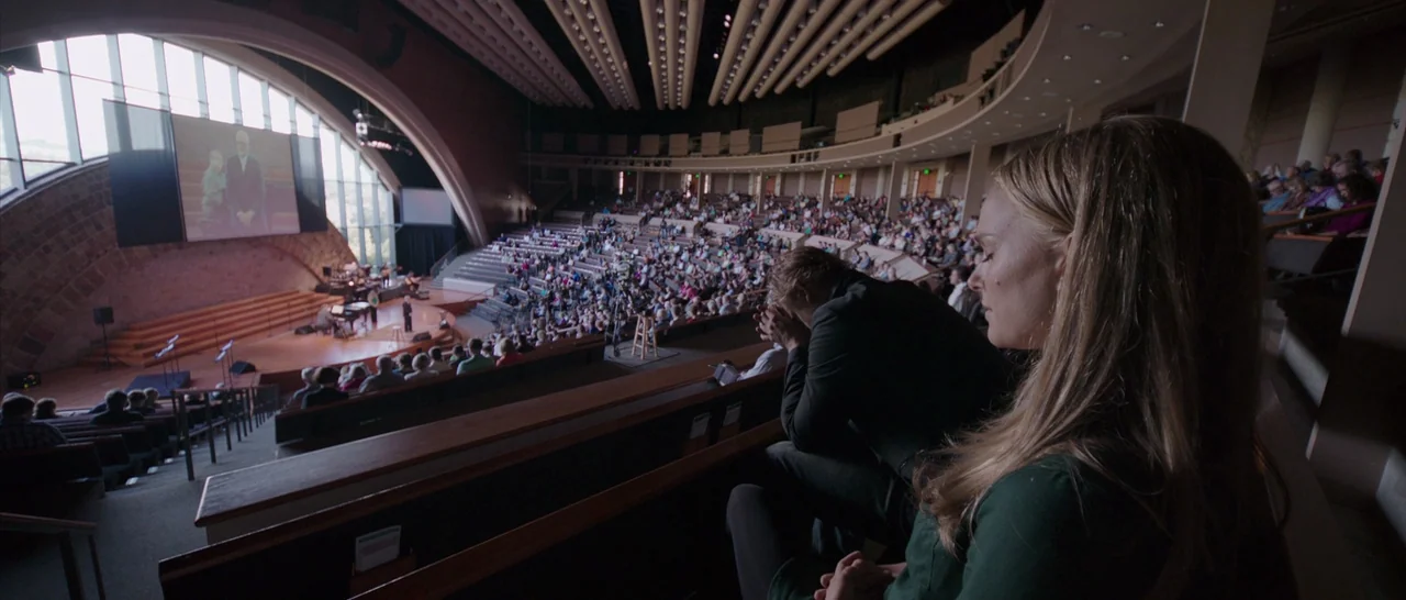 Song to Song (2017) movie still: wide — A modern, large-scale auditorium with tiered seating and a curved glass wall. Two people sit together in the…
