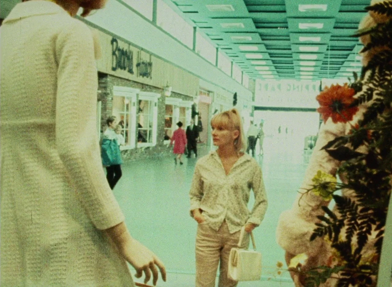 Wanda (1970) movie still: medium — An indoor shopping mall hallway with storefronts lined along a polished light-colored floor. A young woman in…