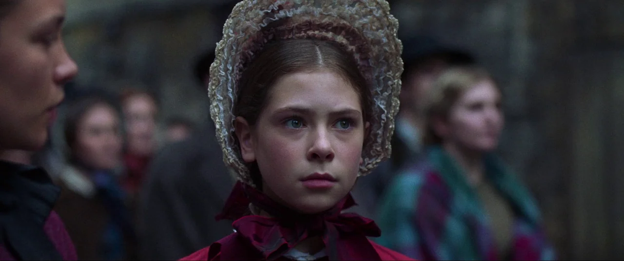 The Wonder (2022) movie still: closeup — An outdoor crowd scene with a young girl centered in the foreground. The girl wears a bonnet and looks…