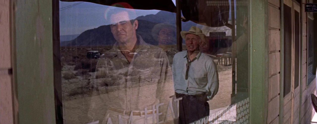 Bad Day At Black Rock (1955) movie still: medium — A wooden building storefront with a large reflective glass window in a desert landscape. Two men…