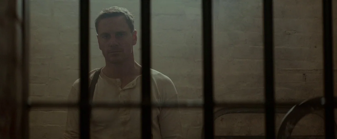 The Light Between Oceans (2016) movie still: medium — A dimly lit room with light-colored brick walls and prominent vertical black metal bars. A man…