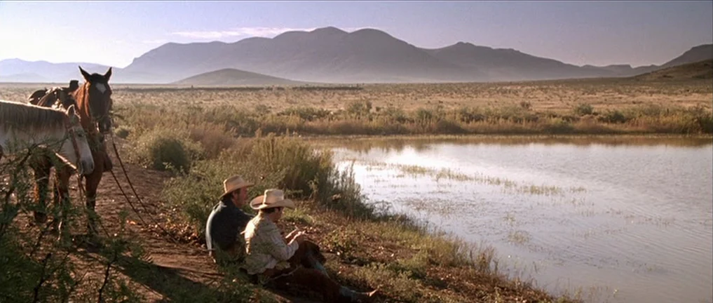 The Three Burials of Melquiades Estrada (2005) movie still: wide — A vast arid landscape features a calm pond reflecting the surrounding hills. Two…