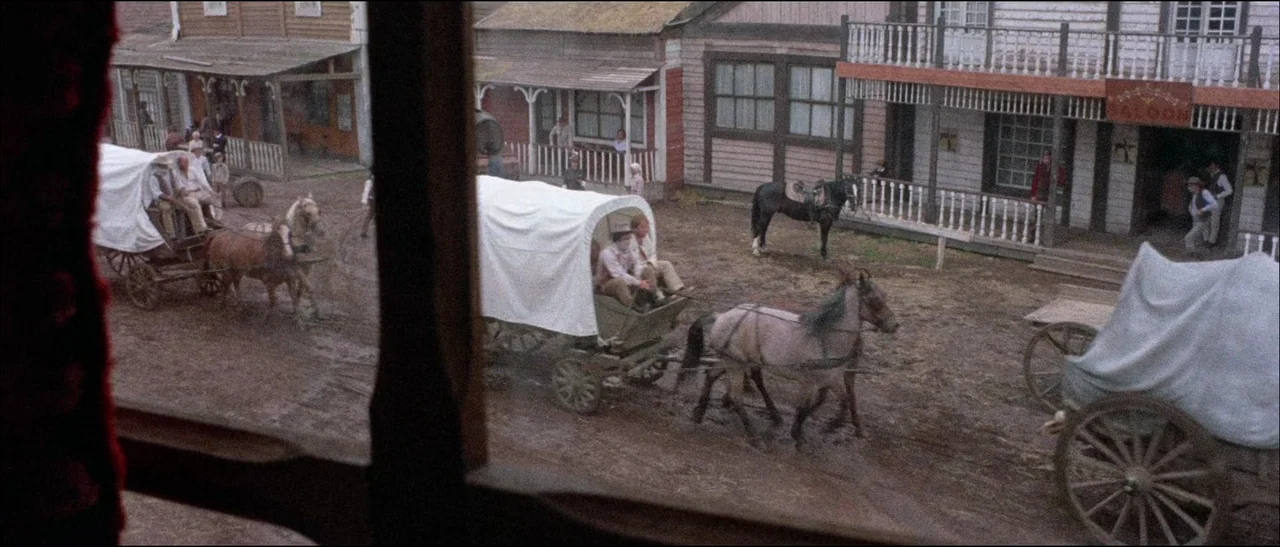 The Grand Duel (1972) movie still: wide — A muddy, underdeveloped street in a western town with several wooden buildings. Covered wagons drawn by…