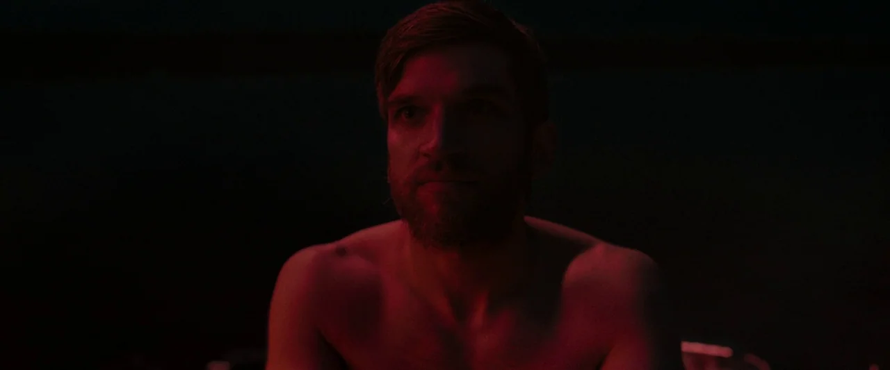 The Night House (2020) movie still: closeup — A man sits shirtless in a dark room illuminated by intense red light. The man stares forward while the…